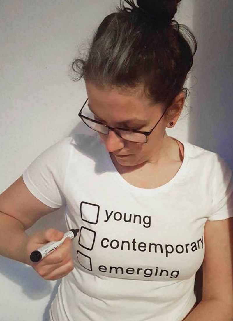 Bild der Künstlerin, die ein weißes T-Shirt mit drei Kästchen und Antwortoptionen drauf an hat. Eine Option streicht sie gerade mit einem Stift an.